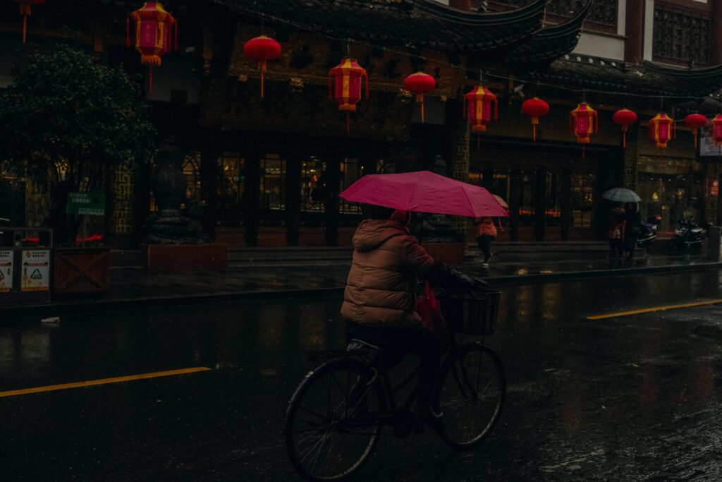 person holding a pink umbrella riding a bicycle in a wet street near a chinese traditional building